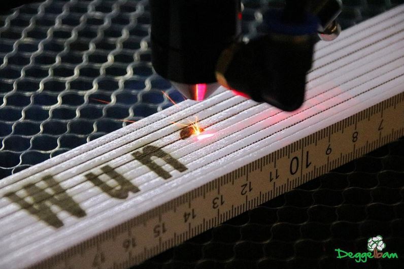 Laser bei der Gravur eines weißen Zollstocks auf einer Wabenplatte.